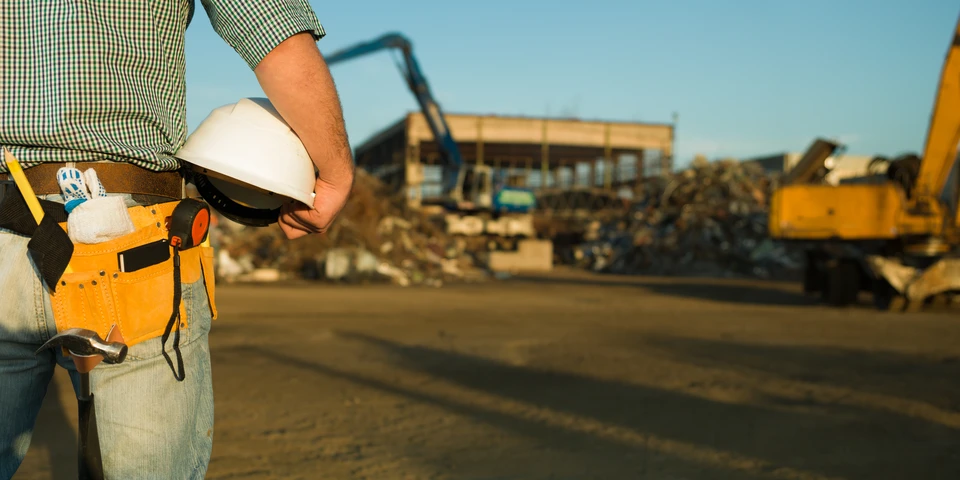 Chantier de construction en ext&eacute;rieur avec ouvrier du b&acirc;timent portant ceinture &agrave; outils orange, casque de s&eacute;curit&eacute; blanc et chemise &agrave; carreaux