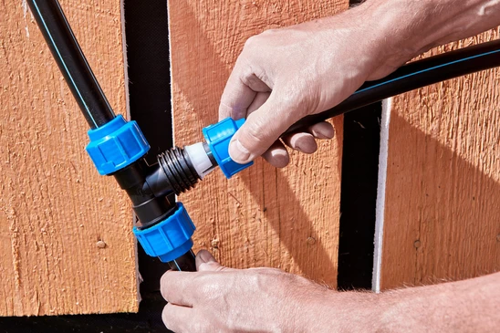 Sc&egrave;ne ext&eacute;rieure, mains d'un technicien assemblant des raccords de plomberie contre une cl&ocirc;ture en bois