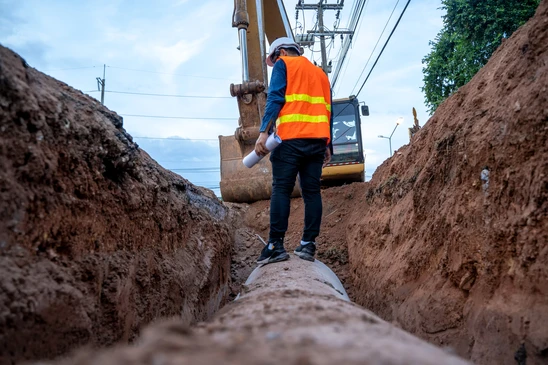 Chantier de travaux publics en ext&eacute;rieur avec ouvrier en gilet haute visibilit&eacute; orange inspectant une canalisation dans une tranch&eacute;e