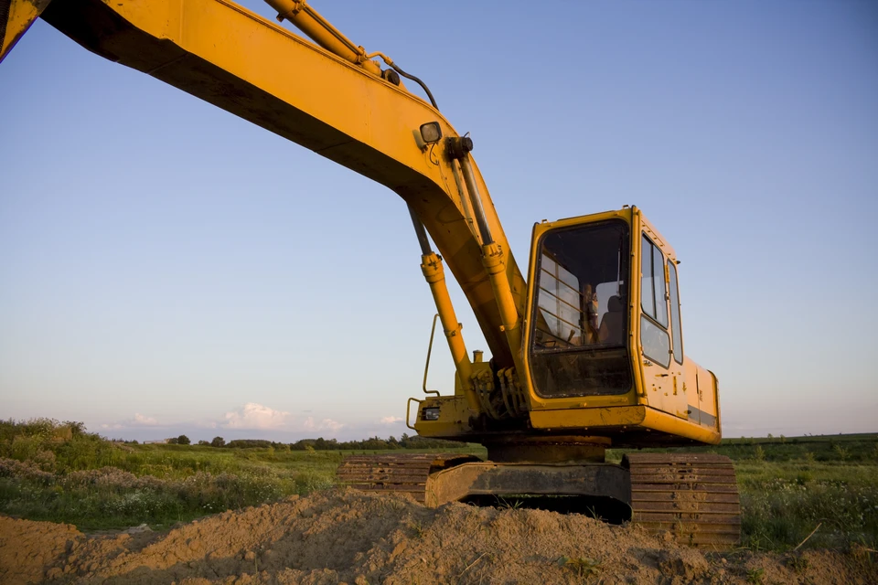 Opérateur en équipement de sécurité orange et casque conduisant un engin de chantier depuis sa cabine vitrée, environnement extérieur avec paysage montagneux en arrière-plan